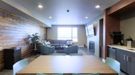 This single-floor space is designed with a clean, contemporary aesthetic that balances comfort and functionality. The open-plan layout seamlessly connects a dining area in the foreground — featuring a light wood table and teal upholstered chairs — with a spacious lounge area beyond. A striking exposed brick accent wall anchors the living space, adding warmth and texture against the smooth, neutral-toned walls and ceilings. The lounge is furnished with a large sectional sofa and coordinating teal armchairs arranged around a central coffee table, creating a welcoming and social atmosphere. A wall-mounted flat-screen television sits above a sleek, modern fireplace, serving as a natural focal point for the seating arrangement. Recessed ceiling lighting throughout provides a bright and even illumination, while a large window allows natural light to fill the room. Built-in wooden shelving and cabinetry along one wall adds both storage and architectural interest. The overall ambiance is warm and inviting, blending industrial elements like the exposed brick with modern finishes to create a versatile, well-appointed communal environment suited for both relaxed gatherings and everyday use.