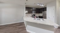 This single-story, one-bedroom, one-bathroom residence offers a well-designed layout that balances functionality and modern style. The kitchen serves as a focal point of the unit, featuring rich dark cabinetry, granite countertops, a mosaic tile backsplash, and a full suite of stainless steel appliances including a range, microwave, and refrigerator. A peninsula with an undermount sink and pendant lighting overhead creates a natural division between the kitchen and the adjoining living area, making it ideal for both everyday use and casual entertaining. Wood-style flooring runs throughout the main living spaces, adding warmth and continuity to the open-concept design.

The bedroom is a comfortable, carpeted retreat equipped with a ceiling fan and a window that allows natural light to filter in while offering views of the surrounding exterior. Neutral wall tones throughout the unit contribute to a calm, versatile ambiance. The bathroom features a granite-topped vanity with dark cabinetry, an undermount sink, and a large mirror, maintaining the consistent design language found throughout the home. The overall layout, as seen in the floor plan, is efficiently arranged to maximize usable space within a compact, single-level footprint.