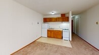 This single-floor unit is efficiently laid out, making practical use of its compact footprint. A hallway serves as the central connector, linking the living space and kitchen zone with clean, straightforward circulation. The living area is finished with wall-to-wall carpet in a neutral beige tone, complemented by crisp white walls and warm wood baseboard trim. A textured ceiling adds a subtle visual detail overhead. Natural light enters through windows fitted with horizontal blinds, brightening the space and creating a pleasant, airy atmosphere. A wall-mounted air conditioning unit and baseboard heating elements provide year-round climate control throughout the carpeted areas.

The kitchen is positioned adjacent to the main living space and transitions from carpet to a smooth vinyl floor. It features a row of warm-toned wood cabinetry above and below the counter, paired with white appliances including a gas range, refrigerator, and a single-basin sink. A ceiling-mounted light fixture provides direct illumination over the kitchen area. The overall palette of the unit is consistent and neutral, with white walls and light finishes creating a clean, understated environment throughout. The hallway, visible from multiple vantage points, connects the spaces cohesively and includes additional built-in storage access.