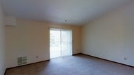 This single-floor residential space offers a clean, straightforward layout designed for comfortable everyday living. The living area is a standout feature, characterized by soft wall-to-wall carpeting that adds warmth underfoot and a neutral color palette across the walls that creates a calm, inviting atmosphere. A large sliding glass door serves as the focal point of the main living area, framed by vertical blinds that allow for flexible light control. The door floods the room with natural light and provides a direct connection to the outdoors, offering views of surrounding greenery. The textured ceiling adds a subtle architectural detail overhead, while a centrally located ventilation vent reflects a practical, functional design approach. Dark baseboards run along the perimeter of the room, providing a subtle contrast to the light-toned walls and carpeting. The overall ambiance of the space is bright, airy, and understated — well-suited for those who value a clean, move-in-ready environment with a simple, functional aesthetic. The single-floor layout ensures easy accessibility throughout, making it a practical and comfortable living option.