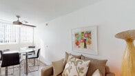 Located at 9500 Zelzah Ave in the Northridge neighborhood of Los Angeles, this single-story unit offers a practical and well-organized layout designed for comfortable living. The combined living and dining area serves as the heart of the space, featuring wood-look flooring throughout, a ceiling fan, and natural light filtering in through vertical blinds. A cozy loveseat anchors the living area alongside a coffee table and vibrant framed artwork, while a glass-top dining table with sleek chrome chairs completes the shared space. The kitchen is efficiently designed with white cabinetry, granite-style countertops, and stainless steel appliances including a refrigerator, range, and microwave — providing all the essentials in a compact footprint. The bedroom accommodates two beds separated by a decorative room divider screen, with a cool gray accent wall adding depth and style. A built-in vanity with a sink and overhead track lighting is integrated into the bedroom, offering added convenience. The bathroom is clean and simply appointed with white finishes, wood-look flooring, and understated décor. The overall ambiance throughout is bright, modern, and functional, with a neutral palette accented by colorful artwork and warm lighting details.