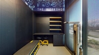 "The Workshop" is a thoughtfully designed bicycle maintenance and parking facility located in Hayward, California. The single-level space is purpose-built to serve cyclists with a comprehensive suite of amenities in a compact, well-organized environment.

The interior is defined by its dark, industrial aesthetic. Floor-to-ceiling pegboard wall panels in matte black line the space, providing versatile tool organization surfaces and giving the area a cohesive, utilitarian character. A bold graphic mural featuring bicycle components and the "The Workshop" signage spans the upper wall and ceiling, reinforcing the space's identity and adding visual interest.

A central built-in workstation includes a dark-surfaced work desk accompanied by rolling mechanic stools, illuminated wood-toned shelving above for organized storage, and lower cabinetry for securing tools and supplies. A yellow mechanic's creeper and a rolling work bench add to the functional equipment available for maintenance tasks.

Adjacent to the workshop area, a dedicated bike parking zone is accessible via a secured entry door equipped with a key-card reader, ensuring controlled access. A frosted glass entry door allows natural light to filter into the space while maintaining privacy. The smooth concrete flooring throughout is durable and easy to maintain, completing the practical, professional design of this well-appointed facility.