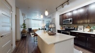 This single-floor residence showcases a thoughtfully designed open-concept layout that seamlessly connects the kitchen, dining, and living areas. The kitchen is a functional centerpiece, featuring rich dark-espresso cabinetry extending to the ceiling, stainless steel appliances including a refrigerator, electric range, and microwave, along with a tile backsplash and generous counter space. A freestanding island with a white quartz countertop and an integrated sink provides additional workspace and casual seating, illuminated by elegant pendant lights overhead. Track lighting along the kitchen ceiling adds targeted, adjustable illumination throughout.

Dark hardwood-style flooring runs consistently through the main living areas, creating a cohesive and warm aesthetic. The living area benefits from a large window that allows abundant natural light to fill the space, enhancing the bright, white walls and airy atmosphere.

The bedroom is comfortably sized, featuring large windows with blinds and sheer curtains, plush carpet flooring, and ample wall space. The adjoining bathroom offers a single-sink vanity with dark cabinetry, a combination tub and shower, and matching hardwood-style flooring, with direct access connecting the bedroom and bath for added convenience.