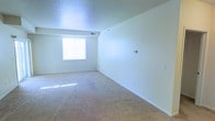 This single-floor space presents a straightforward and functional layout centered around a spacious main living area. The room is finished with wall-to-wall beige carpeting that extends throughout, providing a soft, neutral foundation that complements the clean, white painted walls. A textured popcorn ceiling adds subtle dimension overhead, while a centrally positioned window allows natural light to filter into the room, casting bright patches across the floor and contributing to an open, airy feel. Visible HVAC vents are integrated into both the ceiling and upper wall areas, ensuring climate control throughout the space. White baseboards frame the perimeter of the room, offering a crisp contrast to the carpeted floor. The walls are smooth and unadorned, providing ample flexibility for personalization. Overall, the space maintains a minimal, clean aesthetic with a neutral color palette throughout. The single-floor layout and open configuration make it well-suited for a variety of furniture arrangements, and the abundance of natural light enhances the sense of openness within the main living area.
