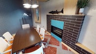 This single-floor meeting room combines functional design with eclectic, characterful details. The centerpiece of the space is a striking floor-to-ceiling fireplace clad in dark brown brick-style tiles, accented by a bold band of turquoise vertical tiles that add a vibrant pop of color. A decorative sculpted fish mounted above the fireplace surround serves as a memorable artistic focal point, lending the room a distinctive personality.

A medium-toned wood conference table runs along one side of the room, surrounded by modern white ergonomic chairs, providing a comfortable and professional setting for meetings or gatherings. Overhead, a cluster of polished chrome pendant lights with flat circular shades delivers focused illumination and a contemporary industrial aesthetic.

The flooring features a layered combination of patterned rugs in warm red and orange tones over a neutral base, adding texture and warmth underfoot. A mirrored nook with a coat rack near the entrance provides practical storage. The walls are finished in cool gray tones, complementing the overall palette. Together, these elements create an inviting, well-appointed meeting environment that balances professionalism with creative, lodge-inspired charm.
