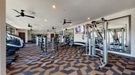 Located at 4144–4148 Belt Line Rd in Addison, TX, this single-floor fitness center offers a spacious and thoughtfully designed workout environment. The space is unified by a bold geometric-patterned carpet in earthy tones of brown, black, and tan, lending a modern and dynamic aesthetic throughout.

The strength training area is generously equipped with a full rack of dumbbells, multiple cable and multi-use weight machines, adjustable benches, and functional training attachments. Full-length mirrors line the walls, enhancing the sense of depth and allowing users to monitor their form. Ceiling fans are mounted throughout, ensuring comfortable airflow during workouts, while recessed lighting keeps the space well-lit.

The cardio zone, positioned along a wall of large arched and grid-paned windows, features a row of treadmills, elliptical machines, a stationary bike, and a rowing machine. These floor-to-ceiling windows flood the area with natural light and provide views of the surrounding Addison streetscape. Wall-mounted flat-screen TVs are present throughout for entertainment during workouts.

A dedicated lounge area near the entrance offers seating with a round coffee table, accent chairs, and a striking copper sculptural wall piece, providing a welcoming transition into the facility. The overall ambiance is clean, modern, and motivating, making this fitness center a comprehensive resource for all levels of exercise.