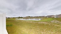This single-story property is situated within a established residential neighborhood, distinguished by its proximity to a community pond that serves as a prominent natural focal point. The exterior setting reveals an expansive grassy landscape that gently slopes toward the water's edge, creating an open and tranquil outdoor environment. The pond itself is a sizable body of water that reflects the surrounding sky, lending a serene, naturalistic quality to the property's backdrop.

The neighborhood context features a row of similarly styled single-story homes with neutral-toned exteriors and pitched rooflines, indicating a cohesive residential development. Young trees are interspersed throughout the community, suggesting the neighborhood is relatively new and continuing to mature. The flat to gently rolling terrain contributes to an open, unobstructed sense of space, with wide sightlines across the pond and surrounding greenery.

The overall ambiance of the setting is quiet and pastoral, well-suited for those seeking a peaceful residential environment with natural water views. The combination of open lawn, pond frontage, and community streetscape provides a balanced blend of natural scenery and suburban accessibility.