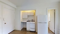 This single-floor space offers a functional and straightforward layout designed to maximize utility. The kitchen is tucked into a dedicated alcove, creating a defined cooking area that keeps the space organized. It is equipped with a gas range and oven, a full-size refrigerator, and a single-basin sink, providing all the essentials for meal preparation. White upper cabinetry lines the top of the alcove, offering ample storage, while under-cabinet lighting adds a warm, practical glow to the workspace. The countertop runs continuously beneath the cabinets, providing sufficient prep space. The flooring throughout features a speckled terrazzo-style surface, adding a classic, durable character to the space. The walls are finished in a clean, bright white, contributing to an open and airy feel. A door adjacent to the kitchen area hints at additional rooms or storage within the unit. Overall, the space presents a no-frills, efficient design that prioritizes functionality, making it well-suited for those seeking a practical and easy-to-maintain living environment.