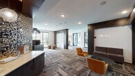 This single-floor amenity space in Frisco, TX, is designed with a contemporary aesthetic that balances functionality and comfort. The open layout is anchored by a distinctive mixed-tone, herringbone-patterned carpet that flows throughout, complementing the neutral walls and dark structural accents that define each zone within the space.

The main lounge area features multiple seating arrangements, including a gray sectional sofa paired with amber accent chairs and a low-profile coffee table, all positioned beneath a wall-mounted flat-screen television. Recessed lighting and dark-trimmed ceiling details add depth and visual interest overhead.

A dedicated workstation area includes desktop computer terminals set along dark cabinetry, accompanied by a stylish pendant chandelier. Nearby, a beverage counter with a textured gold backsplash and illuminated display provides a sophisticated focal point. Curated artwork adorns the walls throughout, contributing to a polished, gallery-like atmosphere.

Private seating nooks with high-back leather chairs and individual side tables offer quieter settings, each framed by large black-trimmed windows that bring in natural light and exterior views. Glass-panel doors connect to adjacent spaces, enhancing the sense of openness. The overall ambiance is modern and welcoming, suited for both social and productive use.
