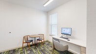 Located at 733 N Drennan St in Houston's East Downtown (EaDo) neighborhood, this compact office space is thoughtfully organized into two distinct areas connected by an open doorway. The layout maximizes functionality within its modest footprint, making it well-suited for focused work or small consultations.

The main room features a built-in white desk workstation equipped with a computer monitor, keyboard, and mouse, accompanied by a modern upholstered chair. Natural light filters in through a window dressed with white horizontal blinds, creating a bright and comfortable work environment. Overhead fluorescent lighting ensures consistent illumination throughout.

An adjoining alcove-style room provides a more intimate setting, furnished with a round wooden table and two mid-century-inspired wooden chairs with padded seats — ideal for small meetings or collaborative discussions.

Throughout both areas, the same bold patterned carpet tiles in shades of gray, charcoal, and vibrant lime green add a contemporary and energetic visual element, contrasting effectively with the clean white walls. The space maintains a clean, modern aesthetic with minimal ornamentation, emphasizing practicality and efficiency. Overall, this well-appointed office space offers a functional and visually dynamic environment suited for professional use.