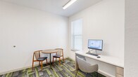 Located at 733 N Drennan St in Houston's East Downtown (EaDo) neighborhood, this compact office space is thoughtfully organized into two distinct areas connected by an open doorway. The layout maximizes functionality within its modest footprint, making it well-suited for focused work or small consultations.

The main room features a built-in white desk workstation equipped with a computer monitor, keyboard, and mouse, accompanied by a modern upholstered chair. Natural light filters in through a window dressed with white horizontal blinds, creating a bright and comfortable work environment. Overhead fluorescent lighting ensures consistent illumination throughout.

An adjoining alcove-style room provides a more intimate setting, furnished with a round wooden table and two mid-century-inspired wooden chairs with padded seats — ideal for small meetings or collaborative discussions.

Throughout both areas, the same bold patterned carpet tiles in shades of gray, charcoal, and vibrant lime green add a contemporary and energetic visual element, contrasting effectively with the clean white walls. The space maintains a clean, modern aesthetic with minimal ornamentation, emphasizing practicality and efficiency. Overall, this well-appointed office space offers a functional and visually dynamic environment suited for professional use.