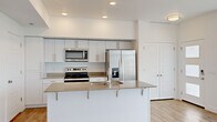 This contemporary two-story residence offers a well-organized layout spread across two floors, with the main living areas on the lower level and the bedrooms above. The open-concept kitchen and living room anchor the first floor, featuring light hardwood-style flooring, recessed lighting, and a clean, neutral color palette throughout. The kitchen is appointed with white shaker-style cabinetry, warm-toned quartz countertops, a central island with a double stainless steel sink and breakfast bar seating, and a full suite of stainless steel appliances including a range, over-the-range microwave, and French door refrigerator with ice maker. The living area flows naturally from the kitchen, offering an open and airy feel. The attached garage provides convenient direct-access parking and additional storage. Upstairs, the upper floor houses both bedrooms, four full bathrooms, and two closets. The generously sized primary bedroom features plush carpeting, windows with outdoor views, and ample natural light. Walk-in closets with built-in rod shelving offer substantial storage capacity. A dedicated in-unit laundry room equipped with a full-size front-loading washer and dryer adds everyday convenience. The home's crisp white walls, modern panel doors, and consistent finishes create a cohesive, move-in-ready environment throughout.