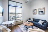 This single-story studio in Jonesboro, Arkansas, offers a thoughtfully designed open-concept layout that maximizes every square foot. The living area is anchored by a large picture window that floods the space with natural light and provides expansive outdoor views, creating an airy atmosphere despite the compact footprint. Light wood-tone flooring runs throughout, contributing to a warm, cohesive aesthetic. A wall-mounted flat-screen TV and a small dining table with modern chairs make efficient use of the living space.

The galley-style kitchen features light wood cabinetry, white quartz countertops, and stainless steel appliances, including a range, microwave, refrigerator, and dishwasher. The tall ceiling height above the kitchen enhances the sense of openness. The sleeping area is partitioned from the main living space, providing a degree of privacy, and is accented by high walls that emphasize the vertical scale of the unit.

The full bathroom includes a shower with a curtain surround, standard fixtures, and light wood flooring consistent with the rest of the unit. A stacked washer and dryer unit is conveniently integrated within the bathroom. The overall design is clean, modern, and functional, making this studio a well-appointed space in the heart of Jonesboro.