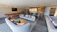 This contemporary amenity lounge is thoughtfully designed to blend social gathering, entertainment, and relaxation into one cohesive space. The open-plan layout is anchored by large-format slate tile flooring and geometric patterned area rugs that define distinct activity zones throughout.

At the heart of the space is a striking wet bar and kitchen area, distinguished by a live-edge wood communal counter atop a tile base, lined with modern bar stools. A row of globe pendant lights cascades above the counter, casting warm ambient light. Reclaimed wood paneling wraps the surrounding walls, adding rustic warmth and texture that contrasts beautifully with the clean white cabinetry and quartz countertops of the prep area.

Adjacent to the bar, a dedicated lounge zone features plush modular sofas accented with vibrant orange and teal throw pillows. A mounted flat-screen television is set against a full reclaimed wood accent wall, with a round marble side table completing the seating arrangement.

The game area includes a full-size shuffleboard table positioned beneath a wall-mounted television, set against white brick walls with built-in accent lighting. Large sliding glass doors and expansive windows flood the space with natural light and open directly onto an outdoor pool deck, seamlessly connecting the interior lounge to the exterior amenities beyond.