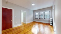 Located at 577 Prospect Ave in West Hartford, CT, this single-floor unit offers a clean and functional layout with classic design elements throughout. Rich, warm hardwood floors span the main living and bedroom areas, reflecting natural light that streams in through multi-pane windows dressed with horizontal blinds, creating a bright and inviting atmosphere. The walls are finished in a soft, neutral gray tone, complemented by crisp white trim and baseboards that give the space a polished, cohesive look. The living area is generously proportioned, offering an open, airy feel with room to arrange furnishings comfortably. The kitchen is efficiently designed, featuring warm-toned wood cabinetry, stone-look tile flooring, a stainless steel gas range with an overhead ventilation hood, and a full-size stainless steel refrigerator. The bathroom is clean and well-appointed, with a tub-shower combination, a white vanity with a single-basin sink, a medicine cabinet with mirror, and neutral stone-look tile flooring. A cast iron radiator is visible in one of the rooms, indicative of the building's classic construction. The entry features a rich, dark wood door, adding warmth and character to the space. Overall, the unit combines timeless architectural details with practical modern updates.