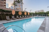 Located in the heart of Downtown Los Angeles at 225 S Grand Ave, this outdoor amenity deck offers a resort-inspired retreat set against the dramatic backdrop of the city skyline. The centerpiece of the space is a generously sized, crystal-clear swimming pool with a dedicated spa, both finished in sleek contemporary tile and surrounded by wide, open pool decking ideal for lounging and socializing. Rows of teak-framed lounge chairs with neutral mesh seatings are neatly arranged poolside, each accompanied by small side tables and accented by distinctive round decorative mirrors that add a modern, artistic touch to the outdoor environment. Lush vertical green walls line the perimeter, softening the urban surroundings and providing a sense of privacy and natural texture throughout the space. Towering palm trees punctuate the landscape, enhancing the California-resort atmosphere. Glass railings along the outer edges preserve unobstructed sightlines to the surrounding high-rise buildings, while stairwell access with stainless steel railings provides convenient connectivity. The overall ambiance blends urban sophistication with a relaxed, sun-soaked aesthetic, making this outdoor deck a standout amenity in the Downtown Los Angeles landscape.