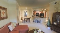 This single-floor residence showcases an open-concept floor plan that seamlessly connects the living, dining, and kitchen areas into one cohesive, flowing space. Warm neutral tones on the walls are complemented by crown molding and arched architectural details, adding a refined touch throughout.

The living area is anchored by a plush, terracotta-toned sectional sofa and a dark wood coffee table, creating a comfortable and welcoming atmosphere. Adjacent to it, the dining area features an elegant dark dining table surrounded by upholstered chairs, with a large decorative mirror enhancing the sense of depth and light.

The kitchen is a functional centerpiece, outfitted with warm wood cabinetry, dark countertops, and black appliances including a refrigerator, microwave, and stove. A curved breakfast bar with bar stools provides additional seating and serves as a natural divider between the kitchen and living spaces. Pendant lighting above the bar and track lighting throughout add both task and ambient illumination. Built-in shelving niches in the walls offer display space, contributing to the home's polished, carefully considered design.