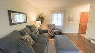 This three-floor home presents a thoughtfully arranged living space designed for both comfort and functionality. The living room, visible in the available imagery, establishes a welcoming tone for the residence. It features warm-toned walls complemented by hardwood flooring, creating a cohesive and grounded aesthetic. Natural light filters into the room through a window dressed with sheer white curtains, brightening the space with a soft, airy quality.

The seating arrangement centers around a plush, gray sectional sofa accented with patterned throw pillows, paired with a matching ottoman that adds both comfort and versatility. A coordinating patterned armchair completes the seating area, providing additional accommodation. Subtle decorative touches, including a potted plant, a table lamp, and framed wall art, contribute to the room's lived-in charm.

Extending across three floors, the home offers generous vertical space and the potential for clearly defined living, sleeping, and utility zones across its levels. The overall ambiance of the residence is warm and residential, with an emphasis on comfortable, practical living enhanced by classic interior detailing.