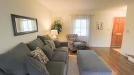 This three-floor home presents a thoughtfully arranged living space designed for both comfort and functionality. The living room, visible in the available imagery, establishes a welcoming tone for the residence. It features warm-toned walls complemented by hardwood flooring, creating a cohesive and grounded aesthetic. Natural light filters into the room through a window dressed with sheer white curtains, brightening the space with a soft, airy quality.

The seating arrangement centers around a plush, gray sectional sofa accented with patterned throw pillows, paired with a matching ottoman that adds both comfort and versatility. A coordinating patterned armchair completes the seating area, providing additional accommodation. Subtle decorative touches, including a potted plant, a table lamp, and framed wall art, contribute to the room's lived-in charm.

Extending across three floors, the home offers generous vertical space and the potential for clearly defined living, sleeping, and utility zones across its levels. The overall ambiance of the residence is warm and residential, with an emphasis on comfortable, practical living enhanced by classic interior detailing.