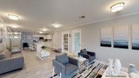 This single-floor space offers a thoughtfully designed open-concept layout that seamlessly connects the living and kitchen areas, creating a welcoming and functional environment. The interior is defined by a consistent coastal-inspired color palette of soft blues, grays, and crisp whites, complemented by crown molding, light wood-look flooring, and flush-mount lighting throughout.

The spacious living area features multiple seating arrangements, including gray upholstered sofas and accent chairs, arranged around a white coffee table atop a patterned area rug. Coastal-themed artwork adorns the walls, reinforcing the calming aesthetic. French doors with glass panes connect the living zones, allowing natural light to flow freely between spaces.

The kitchen is clean and modern, outfitted with white shaker-style cabinetry, light countertops, a stainless steel refrigerator, and a built-in microwave. A central island with bar stool seating provides additional workspace and a casual dining option. A closet pantry adds practical storage.

A secondary lounge area near the main entrance offers additional flexible seating, accented by blue-painted walls, decorative vases, and curated artwork. The front entry features a paneled door and an exit sign, suggesting a multi-use or communal purpose for the space. Overall, the design balances comfort, functionality, and a cohesive modern-coastal aesthetic.