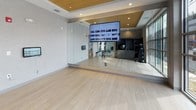 This thoughtfully designed yoga and fitness studio offers a clean, contemporary environment well-suited for a variety of exercise and wellness activities. The space features light-toned hardwood flooring that extends throughout, providing a warm yet polished foundation underfoot. A tray ceiling with a striking wood-paneled inset and recessed lighting adds architectural interest and a sense of refined detail overhead.

Floor-to-ceiling mirrors line one wall, visually expanding the room and supporting movement-based workouts. A large flat-screen display is mounted prominently for guided classes or workout programming. An expansive window along one wall invites abundant natural light and provides views of the surrounding outdoor environment, creating a bright and energizing atmosphere.

Equipment storage is handled by a sleek, dark built-in shelving and cabinet unit that houses stability balls, kettlebells, and stacked medicine balls, keeping the open floor area free and uncluttered. A dumbbell rack with color-coded weights adds a functional and vibrant accent near the entry. A modern door with frosted glass panels provides access to the broader facility, which includes additional cardio and strength training equipment visible through the mirrored walls. The overall ambiance is calm, organized, and purpose-built for focused fitness and wellness practice.