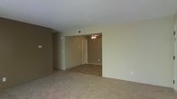 This single-floor space presents a straightforward and functional layout characterized by neutral, earthy tones throughout. The main living area is finished with wall-to-wall carpeting in a warm beige hue, contributing to a comfortable and understated atmosphere. Walls are painted in muted tan and cream tones, creating a cohesive, neutral palette across the space.

An open passage connects the carpeted living area to an adjoining room, which features hard surface tile flooring in a complementary earth-tone pattern — offering a practical contrast to the softer carpeted areas. This secondary room is equipped with a ceiling fan and light fixture, providing both ventilation and ambient lighting. A white paneled door leads further into the space, suggesting additional rooms or storage beyond.

Overhead, a standard white ceiling features a visible HVAC vent, indicating a central air system. Subtle architectural details, such as the angled wall transition between areas, add modest visual interest to the otherwise clean and simple design. The overall ambiance is calm and move-in ready, suited for a variety of uses and personal styling preferences.