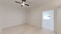 This single-floor space in Greenville, SC, showcases a contemporary design characterized by a crisp, all-white palette, light wood-tone flooring, and an open, airy atmosphere. The main living area makes an immediate impression with dramatically tall ceilings, whitewashed exposed brick walls, and large black-framed windows that invite natural light. A bold black entry door adds industrial contrast to the bright interior, and an oversized ceiling fan serves as a functional focal point overhead.

The kitchen is well-appointed with white shaker-style cabinetry extending to impressive heights, dark granite countertops, a black subway tile backsplash, and a full suite of stainless steel appliances, including a range, dishwasher, microwave, and refrigerator. The vertical cabinet layout maximizes storage while emphasizing the room's notable ceiling height.

The bedroom areas feature the same light hardwood flooring and are accented by wainscoting panels along the lower walls, adding architectural detail. Ceiling fans are installed in each room for comfort.

The bathroom is finished in a clean, monochromatic style with glossy white tile flooring, a floating vanity with a dark countertop, a round mirror, and a tiled walk-in shower with black fixtures. Throughout, the space maintains a cohesive modern-industrial aesthetic.