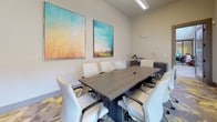 This single-floor conference room offers a polished and professional environment designed to support productive meetings and collaborative work. At the center of the space sits a substantial dark wood-toned boardroom table surrounded by sleek white high-back ergonomic chairs, providing comfortable seating for a full team. A large wall-mounted flat-screen display is positioned at one end of the room, complemented by a dark wood credenza beneath it that offers additional storage and workspace for equipment such as a printer and laptop.

The room is adorned with two large, vibrant abstract paintings featuring warm and cool tones that add a creative energy to the otherwise clean, neutral aesthetic of the light gray walls. Overhead, linear LED lighting fixtures provide bright, even illumination throughout the space.

Natural light filters in through windows fitted with roller shades along one wall, allowing for light control during presentations while maintaining a connection to the outside. The flooring features a distinctive patterned carpet in shades of gray, yellow, and purple, adding visual interest and a contemporary design touch underfoot.

A framed glass-panel door connects the conference room to an adjacent open area, ensuring easy access while maintaining an enclosed, focused atmosphere. The overall ambiance is modern, clean, and functional — well-suited for business meetings, presentations, or team discussions.