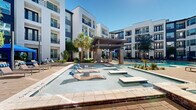 This contemporary multi-story residential complex showcases a striking exterior defined by a bold combination of navy blue horizontal siding, crisp white stucco, and dark-framed windows and balconies. Spanning two floors, the building wraps around a beautifully designed courtyard that serves as the centerpiece of the community.

The outdoor amenity area is anchored by a generously sized resort-style swimming pool with crystal-clear water, complemented by a separate spa and shallow wading areas fitted with built-in lounge ledges. Surrounding the pool is an expansive paver deck appointed with rows of cushioned chaise lounges, shaded blue canopy cabanas, and a pergola-covered seating and dining area — ideal for both relaxation and outdoor entertaining.

Thoughtfully curated landscaping integrates palm trees, ornamental shrubs, flowering groundcover, and mature shade trees throughout the courtyard, creating a lush, tropical ambiance. Paved walkways wind through the greenery, connecting different zones of the outdoor space and leading to ground-floor unit entrances framed by well-maintained garden beds.

Each level of the building features private balconies that overlook the courtyard, offering residents elevated views of the pool and greenery below. The overall design balances modern architectural aesthetics with resort-inspired outdoor living, creating an inviting and vibrant communal environment.