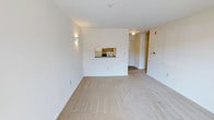 This single-floor unit presents a clean, functional layout centered around a spacious open living area. The room is finished with neutral-toned wall-to-wall carpeting that runs throughout, contributing to a quiet, understated atmosphere. Crisp white walls are kept simple and unadorned, providing a blank canvas that maximizes the sense of openness throughout the space.

A defining architectural detail is the pass-through window connecting the living area to the kitchen, offering a convenient and interactive link between the two spaces. The kitchen, visible through the pass-through, is equipped with standard appliances including a refrigerator and range, fitted within a compact but practical layout.

Wall-mounted sconce lighting adds subtle accent illumination to the living area, complementing the ceiling-mounted fixtures that provide general lighting throughout. A centrally located hallway leads from the living space toward additional rooms, suggesting a straightforward and efficient floor plan. The entry door is positioned along the same wall, keeping circulation logical and unobstructed.

Overall, the space offers a modest, move-in-ready environment with a neutral palette and practical features suited for comfortable everyday living.