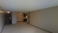 This single-floor space offers a straightforward and functional layout designed to maximize usability. The living area is finished with wall-to-wall carpeting in a neutral tone, complemented by beige walls that create a clean, understated atmosphere throughout. The open floor plan allows the living space to flow naturally toward the kitchen area, which is visible from the main room and features dark espresso-toned cabinetry, a sink, and white appliances. A ceiling light fixture with multiple bulbs illuminates the kitchen and adjoining space, providing adequate overhead lighting. The kitchen also includes a compact refrigerator and what appears to be additional counter space. A textured white ceiling runs throughout the unit, adding a subtle visual element overhead. Dark baseboards provide a grounding contrast against the lighter walls and flooring. The overall ambiance is modest and move-in ready, with a neutral palette that offers flexibility for personalization. The open connection between the living and kitchen areas makes the space feel cohesive and practical for everyday living.