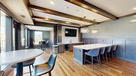 Located in the heart of Saint Paul, this single-floor communal amenity space offers a refined blend of modern design and warm, inviting finishes. The interior lounge is anchored by rich hardwood flooring and striking exposed wooden ceiling beams that lend a sense of architectural character throughout. A fully equipped kitchenette lines one wall, featuring dark wood cabinetry, white quartz countertops, a beverage refrigerator, a sink, and a geometric patterned tile backsplash — all framed by a wall-mounted flat-screen TV overhead. Elegant pendant lighting illuminates a central island with seating, creating a natural gathering point for socializing. Wainscoting in a muted sage-green palette adds a sophisticated, layered look to the walls. A windowed nook area with high-top tables and bar stools offers views of the surrounding cityscape, while French doors open to a spacious rooftop terrace. The outdoor deck is thoughtfully appointed with Adirondack chairs surrounding a fire pit, an outdoor dining table, a gas grill, and an exterior television — all set against panoramic views of downtown Saint Paul. This versatile space is ideal for hosting events or relaxing in an upscale urban setting.