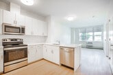 This single-floor unit on Park Drive in Wyandanch, New York, presents a clean, modern aesthetic defined by bright white walls, light hardwood flooring, and an open, airy layout. The kitchen serves as a central feature, equipped with a full suite of stainless steel appliances including a refrigerator, electric range, over-the-range microwave, and dishwasher. White cabinetry extends to the ceiling, maximizing storage, while light countertops and a peninsula with a stainless steel sink create a seamless transition into the open living area. The living space is generously proportioned, benefiting from a large window that invites natural light and offers outdoor views. A dedicated bedroom continues the bright, minimalist feel, with its own window providing additional natural light and a baseboard heating system. The bathroom is finished with a neutral-toned vanity, a framed mirror, hexagonal tile flooring, and an accessible roll-in shower equipped with grab bars and a handheld showerhead. Throughout the unit, flush-mount ceiling lighting and consistent flooring unify the space, creating a cohesive and functional living environment well-suited for comfortable, modern living.