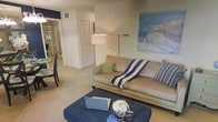 This single-floor residence offers a thoughtfully arranged living space with a warm, coastal-inspired aesthetic. The living room serves as the heart of the home, featuring plush carpeted flooring and neutral wall tones that create a calm, inviting atmosphere. A generously sized sofa in a soft beige tone is accented with blue and white striped throws and patterned pillows, complementing the large coastal landscape painting that anchors the main wall. A deep navy blue ottoman acts as a functional centerpiece, doubling as a coffee table. A sleek floor lamp provides ambient lighting, adding to the room's relaxed yet polished feel. A louvered bifold closet door is visible, offering practical storage seamlessly integrated into the space. The open layout allows for a natural flow between the living area and adjacent spaces, with a glimpse of a dining area to one side. The consistent use of blue and neutral tones throughout ties the space together cohesively, evoking a serene, seaside ambiance. Overall, this residence presents a clean, comfortable, and stylishly understated environment well-suited for everyday living.