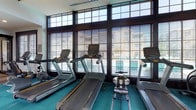 This fitness center is thoughtfully designed to support a full range of workout routines across multiple distinct areas. The main workout floor makes a strong visual impression with its bold, geometric patterned flooring in teal, gray, and yellow tones, complemented by soaring ceilings with exposed black structural beams and ceiling fans. Large floor-to-ceiling windows along the perimeter flood the space with natural light and offer open outdoor views, while venetian blinds allow for adjustable light control. This area is outfitted with a comprehensive selection of strength and cardio equipment, including cable machines, a multi-function weight rack, and a row of cardio machines. A wall of mirrors expands the sense of space and supports proper form during workouts. A dedicated cardio room, positioned along an exterior wall of expansive windows, houses multiple treadmills and a rowing machine, creating a bright and energizing environment for cardio training. A separate studio room provides a quieter space for stretching and functional training, equipped with exercise mats, stability balls, and a wall-mounted screen for guided workout sessions. The entry area features open storage cubbies and leads conveniently into each zone, giving the facility a clean, organized flow throughout.