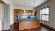 Located on North High Street in Columbus, Ohio's vibrant Short North neighborhood, this single-floor unit offers a thoughtfully designed layout with contemporary finishes throughout. The kitchen is a standout feature, showcasing warm-toned wood cabinetry, dark granite countertops, a glass tile backsplash, and a full suite of stainless steel appliances including a French door refrigerator, electric range, and built-in microwave. A center island with an integrated sink provides additional prep space and casual seating, while rich hardwood flooring and pendant lighting complete the modern aesthetic. Track lighting runs the length of the ceiling, adding both function and style. The open living area continues the hardwood flooring and is bathed in natural light through large windows with slatted blinds, offering views of the surrounding neighborhood. The bedroom features plush wall-to-wall carpeting and a pair of windows that bring in ample daylight. The bathroom is well-appointed with a large walk-in glass-enclosed shower clad in dark tile, a vanity with stone countertop, and clean tile flooring. The overall ambiance is modern and cohesive, with neutral tones, gray trim accents, and quality materials tying the space together throughout.