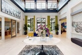 Located at 11181 W 35th Way in Hialeah, FL, this elegantly designed lobby space makes a striking first impression through its dramatic double-height ceilings and expansive black-framed windows adorned with chartreuse grommet curtains. Natural light floods the interior, highlighting the large-format, light-toned tile flooring that runs throughout the open layout.

A central round pedestal table serves as the focal point of the reception area, topped with vibrant floral arrangements that add a welcoming touch. Flanking this centerpiece are carefully curated seating vignettes, including a bold cobalt-blue sofa paired with mustard yellow accent chairs and a dark stone coffee table, as well as plush green velvet armchairs that introduce warmth and texture.

The walls display notable artwork, including a large-scale marble-effect panel and a bold canvas piece, contributing to the space's gallery-like aesthetic. Tall potted fiddle-leaf fig trees punctuate the corners, reinforcing a fresh, biophilic design sensibility. A clerestory-level row of windows at the ceiling's upper tier draws in additional daylight, enhancing the sense of openness. Adjacent rooms are accessible through glass-paneled interior doors, offering seamless connectivity throughout the facility.