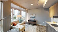 Located in the heart of Seattle's Belltown neighborhood, this contemporary residence offers a thoughtfully designed single-floor layout with a cohesive modern aesthetic throughout. The living area is anchored by large corner windows that frame sweeping city skyline views, flooding the space with natural light. Wood-look flooring, exposed concrete ceiling elements, and track lighting lend an urban-industrial character that complements the warm, neutral color palette.

The kitchen is efficiently laid out and fully equipped with stainless steel appliances, including a refrigerator, range, microwave, and dishwasher. Dark wood cabinetry with high-gloss upper cabinets, white subway tile backsplash, and clean white countertops create a polished, contemporary look. A dining surface adjacent to the kitchen provides a convenient space for casual meals.

The bedroom is a well-appointed retreat featuring a window with city views, warm-toned hardwood floors, and a dedicated workspace with a wood desk. Exposed concrete beam details echo the industrial design elements found throughout the residence.

The bathroom offers a clean, modern finish with a floating vanity, white countertop, large mirror, and a tiled walk-in shower, completing this well-rounded urban living space.