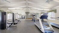 Located at 6470 Timber Bluff Point in Colorado Springs, Colorado, this single-floor community fitness center offers a comprehensive range of workout facilities designed to support a variety of exercise routines. The main gym area is defined by soaring vaulted ceilings with exposed white beams, ceiling fans, and bright fluorescent lighting that creates an open, energizing atmosphere. Durable rubber flooring runs throughout, providing a safe and functional surface for all types of exercise. The cardio section is well-appointed with multiple treadmills, stair climbers, and a stationary bike, complemented by wall-mounted mirrors that enhance the sense of space. A mounted flat-screen television adds convenience for users during cardio sessions. The strength training zone features a selection of cable machines, weight benches, and free weights, all positioned beneath naturally lit windows dressed with decorative valances. A glass-enclosed studio area provides a dedicated space for group fitness or supplemental training with stability balls and additional equipment. Adjoining the main gym is a full racquetball court, finished with a classic hardwood floor marked with regulation court lines and enclosed by bright white paneled walls, offering a spirited option for competitive or recreational play.