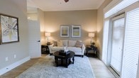 This two-story home exudes a refined yet welcoming character, thoughtfully designed with a cohesive aesthetic that carries throughout the space. The living area serves as a standout feature, showcasing warm beige walls that create a cozy, neutral backdrop complemented by light hardwood-style flooring. Crown molding adds an elegant architectural detail, lending a polished finish to the room. A ceiling fan provides both style and function overhead, while large windows fitted with blinds allow for generous natural light to fill the space, enhancing its open and airy feel. The living room is well-proportioned, offering ample room for comfortable furnishings, including a plush seating arrangement anchored by a soft area rug and a classic round coffee table. Tasteful wall art and coordinated accent lighting contribute to the room's warm ambiance. The two-story layout provides distinct separation between living and private spaces, adding depth and versatility to the overall floor plan. Whether used as a primary residence or otherwise, this home balances practical design with understated elegance, creating an environment that feels both livable and thoughtfully curated from the moment you step inside.