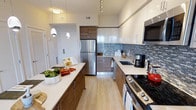 This contemporary single-floor residence showcases a thoughtfully designed interior with clean lines and modern finishes. The kitchen is a standout feature, boasting a galley-style layout that maximizes functionality and style. Sleek, two-tone cabinetry combines high-gloss white upper cabinets with warm wood-grain lower cabinets, creating a visually dynamic and inviting space. A mosaic tile backsplash in cool blue and grey tones adds texture and visual interest, complementing the crisp white countertops throughout. Stainless steel appliances, including a full-size refrigerator, a slide-in range, and an over-the-range microwave, provide a polished, professional-grade cooking environment. Track lighting along the ceiling, paired with elegant pendant lights, ensures the kitchen is well-lit and functional. Light wood-tone flooring runs throughout, adding warmth and cohesion to the space. A generous kitchen island or peninsula offers additional prep space and casual dining potential. The overall ambiance is modern, bright, and efficient, reflecting a well-curated design sensibility ideal for both everyday living and entertaining.