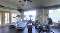 This single-story fitness room offers a well-equipped exercise space designed to meet a variety of cardiovascular training needs. The room houses multiple treadmills along one wall, providing ample options for walking or running workouts. Alongside these, two elliptical cross-trainers and a stair climber offer low-impact alternatives for full-body conditioning.

Natural light flows into the space through multiple windows fitted with horizontal blinds, creating a bright and energizing atmosphere. The walls are painted in a neutral tone accented by a teal border near the ceiling, adding a subtle decorative touch. A ceiling fan with an integrated light fixture helps maintain air circulation throughout the room, enhancing comfort during workouts.

A wall-mounted flat-screen television is positioned for easy viewing from the cardio equipment, allowing users to stay entertained during their sessions. The flooring appears to be durable, dark-toned material suited for high-traffic fitness use. Overall, the space is efficiently arranged to maximize usability while maintaining an open, functional feel.