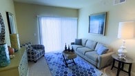 This single-floor residential space offers a comfortable and practical layout designed for everyday living. The living area serves as the heart of the home, highlighted by a large sliding glass door fitted with vertical blinds that allow for adjustable natural light and easy access to the outdoors. The room is finished with plush wall-to-wall carpeting, adding warmth and comfort underfoot. The space is furnished with a generous sofa accented with coordinating throw pillows, a patterned accent chair, and a geometric-print area rug in shades of blue, lending the room a cohesive and inviting aesthetic. A low-profile coffee table with a decorative tray serves as the central focal point of the seating arrangement. Framed wall art adds a finishing touch to the neutral, light-toned walls, contributing to a calm and understated ambiance throughout. The overall layout is efficient and well-proportioned, offering a functional living environment with ample natural light. The single-floor design ensures easy accessibility and a seamless flow throughout the space, making it a practical and comfortable place to call home.