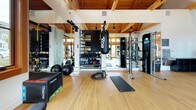 This fitness center at Haven Apartments at Union Station offers a thoughtfully designed workout environment that combines rustic architectural charm with modern amenities. The space is defined by its striking vaulted ceiling with exposed wood beams and tongue-and-groove paneling, which lends warmth and character to the interior. Large wood-framed windows and glass doors flood the facility with natural light while offering views of the surrounding greenery.

The floor is finished in smooth hardwood, providing a clean and durable surface throughout. Floor-to-ceiling mirrors line the walls, enhancing the sense of spaciousness and supporting proper form during workouts. The equipment selection is extensive and varied: a cable resistance machine, TRX suspension trainers, a heavy punching bag, battle ropes, medicine balls, a plyometric box, and a well-stocked dumbbell rack cater to functional and strength training. Cardio options include treadmills and a stationary bike. An adjustable weight bench and exercise mats round out the offerings.

A built-in water bottle filling station and drinking fountain provide convenient hydration. A wall-mounted television and a welcoming entry with a branded area rug complete the experience. French doors connect the fitness center to an adjacent common lounge area, making this a fully integrated amenity space.