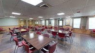 This single-floor community space is designed to accommodate group dining and social gatherings in a comfortable, organized setting. The room features a drop ceiling with recessed rectangular lighting panels that provide bright, even illumination throughout the space. Large windows along the perimeter allow natural light to filter in, contributing to the open and airy feel of the room.

The dining area is furnished with a generous arrangement of rectangular wood-tone tables, each accompanied by cushioned chairs in a deep burgundy red with silver metal frames, offering both style and comfort. The tables are arranged in a spacious layout that allows for easy movement and accessibility throughout the room. Durable wood-look flooring runs throughout, adding warmth to the overall aesthetic.

Colorful decorative touches, including hanging floral ornaments and framed wall art, lend a cheerful and inviting character to the space. A two-tone wall treatment — warm tan on the lower half and neutral tones above — adds visual interest and a sense of structure to the room. An exit door and emergency signage are clearly visible, reflecting attention to safety. Overall, the space exudes a functional yet friendly ambiance well-suited for communal meals, events, or social activities.