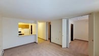 This single-floor space offers a practical, open layout designed for comfortable everyday living. The main living area is finished with wall-to-wall carpeting in a neutral tone, creating a warm and inviting atmosphere. A pass-through counter connects the living space to the kitchen, which features honey-toned wood cabinetry, a white refrigerator, and under-cabinet lighting that adds a soft ambient glow. The kitchen's layout is compact yet functional, making efficient use of available space.

From the main living area, a central hallway provides access to additional rooms, with multiple doorways visible leading to what appear to be bedrooms or utility spaces. Bifold closet doors are a notable feature, offering organized storage solutions along the hallway. The space is finished with consistent white walls and neutral baseboards throughout, contributing to a clean, cohesive look. Overhead lighting keeps the interior well-illuminated. The overall design emphasizes simplicity and functionality, with a straightforward floor plan that connects all areas of the unit in a logical and accessible manner.