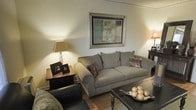 This single-floor space features a thoughtfully arranged living area that exudes a warm and inviting ambiance. The living room centers around a generously sized, neutral-toned sofa accented with decorative throw pillows in earthy tones, providing a comfortable and stylish focal point. Hardwood flooring runs throughout, adding a sense of warmth and classic character to the space. Soft, ambient lighting is provided by table lamps positioned on either side of the room, casting a gentle glow that enhances the cozy atmosphere. A framed piece of abstract artwork with muted blue and grey tones anchors the main wall, adding a refined decorative touch. A dark wood side table sits adjacent to the sofa, offering practical surface space, while a coffee table in the foreground displays a decorative tray. The overall color palette consists of soft whites, warm beiges, and rich wood tones, creating a cohesive and harmonious aesthetic throughout. The layout is efficient yet comfortable, making excellent use of the available space to create a functional and welcoming living environment.