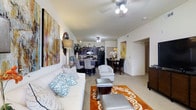 This single-story residence offers an open-concept floor plan that seamlessly integrates the living, dining, and kitchen areas into one cohesive space. The living area is anchored by a plush white sofa accented with decorative teal and white pillows, complemented by a silver ottoman and a warm orange-and-white area rug that adds a bold focal point to the room. A large abstract artwork adorns the wall, lending an artistic touch to the space. Ceiling fans provide both lighting and airflow throughout the main living area and kitchen.

The kitchen features dark cabinetry paired with black appliances, creating a sleek, modern contrast against the lighter walls and flooring. A breakfast bar with bar-height seating transitions smoothly into the dining area, which accommodates a full dining table and chairs, ideal for everyday meals or entertaining.

Neutral-toned carpet and light-colored walls keep the space feeling bright and open throughout. A hallway visible in the background suggests additional private rooms beyond the main living area. Overall, the residence balances a contemporary aesthetic with comfortable, functional furnishings.