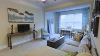 This single-floor living space offers a comfortable and well-arranged interior highlighted by a soft, neutral color palette throughout. The main living area is anchored by plush carpeted flooring and features crown molding along the ceiling, adding a refined architectural detail to the room. A ceiling fan provides both function and a casual aesthetic overhead.

The living area is thoughtfully furnished with a generous sofa dressed in a classic checkered pattern, complemented by a dark wood storage trunk that serves as a coffee table. A cushioned accent chair with floral detailing adds a cozy touch near the window area. The flat-screen television is mounted on a slim, open console table, keeping the space feeling light and uncluttered.

Natural light flows generously into the space through a large window dressed with white horizontal blinds and patterned blue curtains, creating a bright and airy atmosphere. A wooden writing desk and side table are positioned near the window, making excellent use of the natural light. Decorative wall art and carefully chosen accessories throughout the room contribute to a warm, inviting ambiance that balances everyday practicality with personal style.
