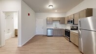 This single-floor, one-bedroom, one-bathroom unit offers a thoughtfully arranged layout that maximizes its compact footprint. Light wood-style flooring runs consistently throughout the space, lending a clean and cohesive aesthetic. Neutral wall tones and flush-mount ceiling lighting contribute to a bright, modern interior.

The kitchen is efficiently designed with taupe-toned cabinetry, light stone-patterned countertops, and a full suite of stainless steel appliances, including a refrigerator, electric range, microwave, and dishwasher. A dedicated pantry closet with built-in shelving sits adjacent to the kitchen, providing notable storage capacity. The kitchen opens directly into the living area, which benefits from a sliding glass door or large window offering outdoor views and natural light.

The bedroom is a well-proportioned private space featuring a window with blinds and a sliding closet with wire shelving. The bathroom includes a wood-toned vanity with a single sink, a lighted mirror, and light-toned flooring. A hallway connects the main rooms efficiently, and the entrance provides a clear transition into the living and kitchen areas.